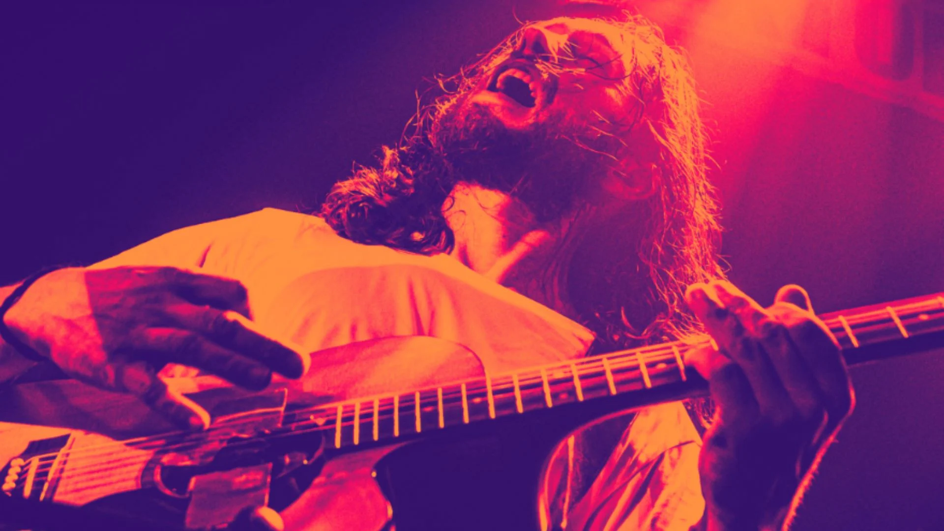 A guitarist with long hair passionately plays an electric guitar on stage, head tilted back and mouth open, bathed in dramatic red and orange lighting.