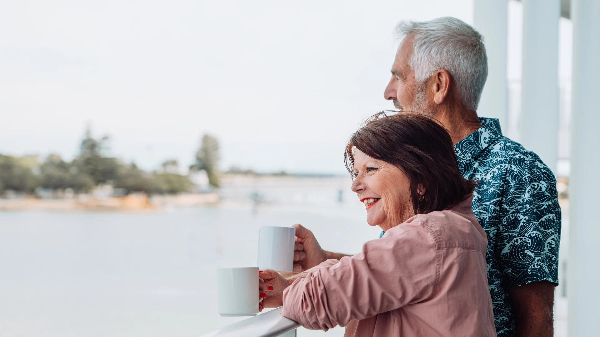 Older couple smiling and holding mugs while looking out at a peaceful waterfront view. They are standing on a balcony, dressed casually, and enjoying a relaxed moment together.
