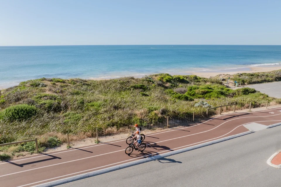 Two cyclists ride along a paved path beside the beach on a sunny day. The ocean is calm and the sky is clear. Vegetation lines the dunes between the path and the beach. A parking lot is visible in the distance.