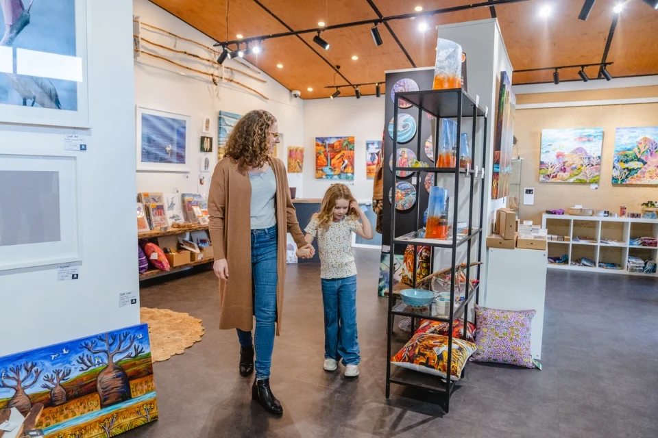 A woman and a child hold hands while walking through an art gallery. The gallery features colorful paintings on the walls and various art pieces on shelves. The setting is warm and well-lit, with wooden ceilings and spotlights.