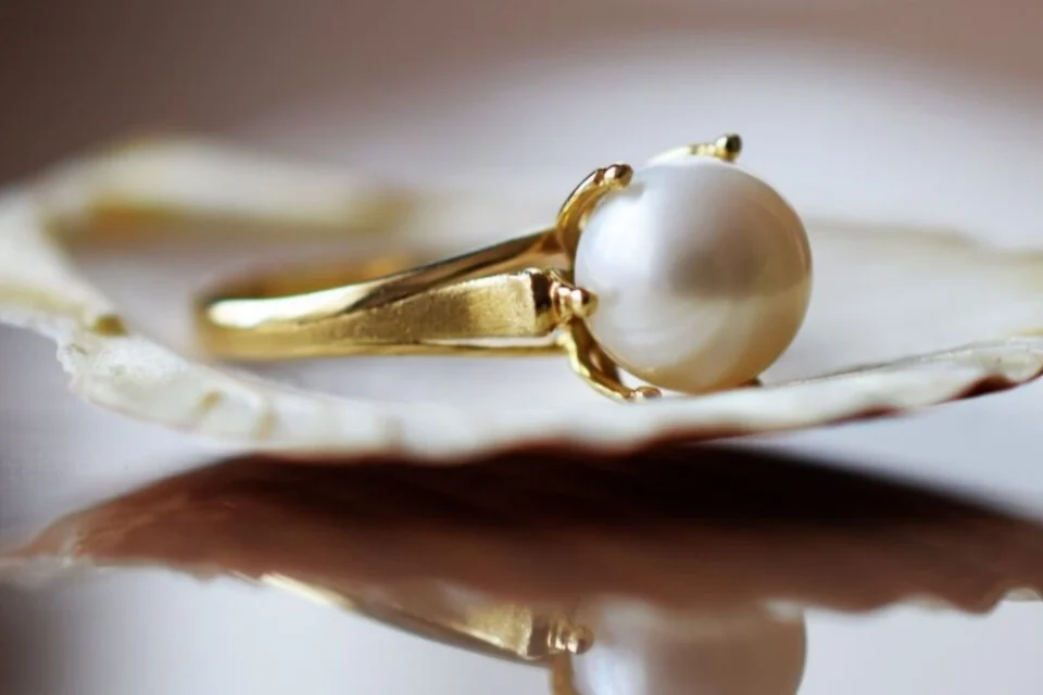 A close-up of a gold ring with a large white pearl set in a prong setting, resting on a soft-focus light background. The pearl is smooth and lustrous, complementing the polished gold band.