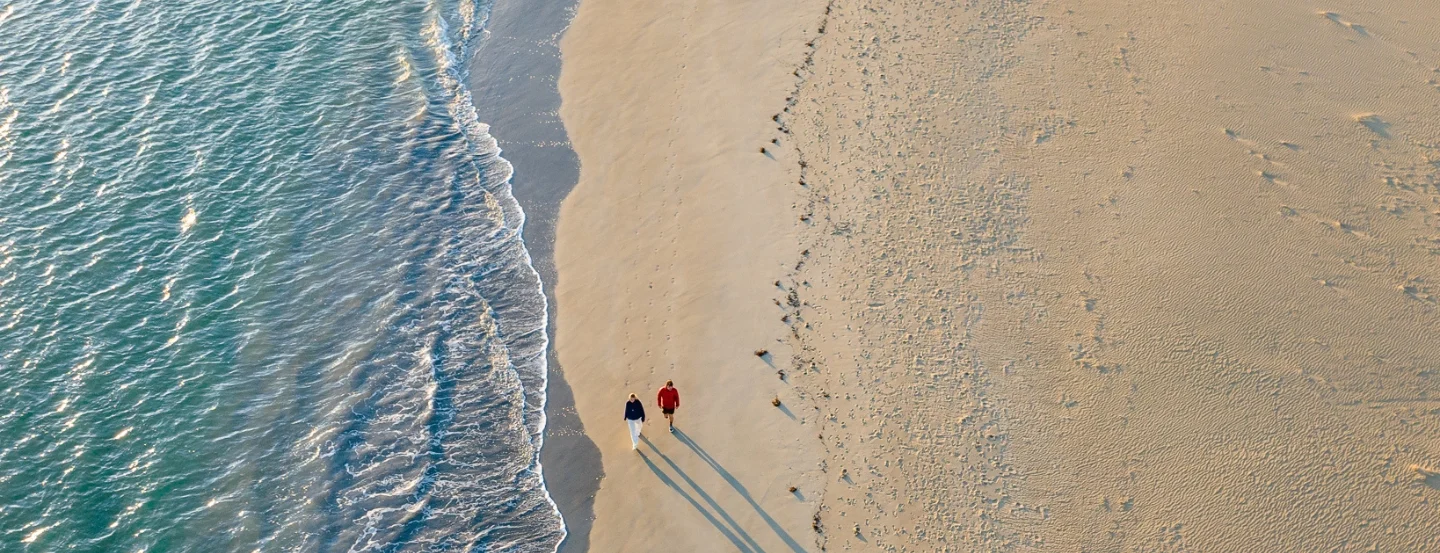 Aerial view of two people walking on a wide, sandy beach. The shoreline curves gently, with waves lapping at the sand on the left. The beach is mostly empty, with the shadows of the people stretching out on the sand.