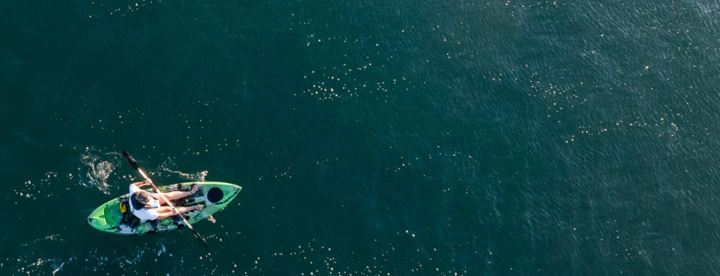 Aerial view of a person kayaking on calm, dark blue water. The kayaker, wearing a hat, is in a green kayak, paddling towards the left of the image, with small ripples trailing behind. The water is dotted with patches of sunlight.