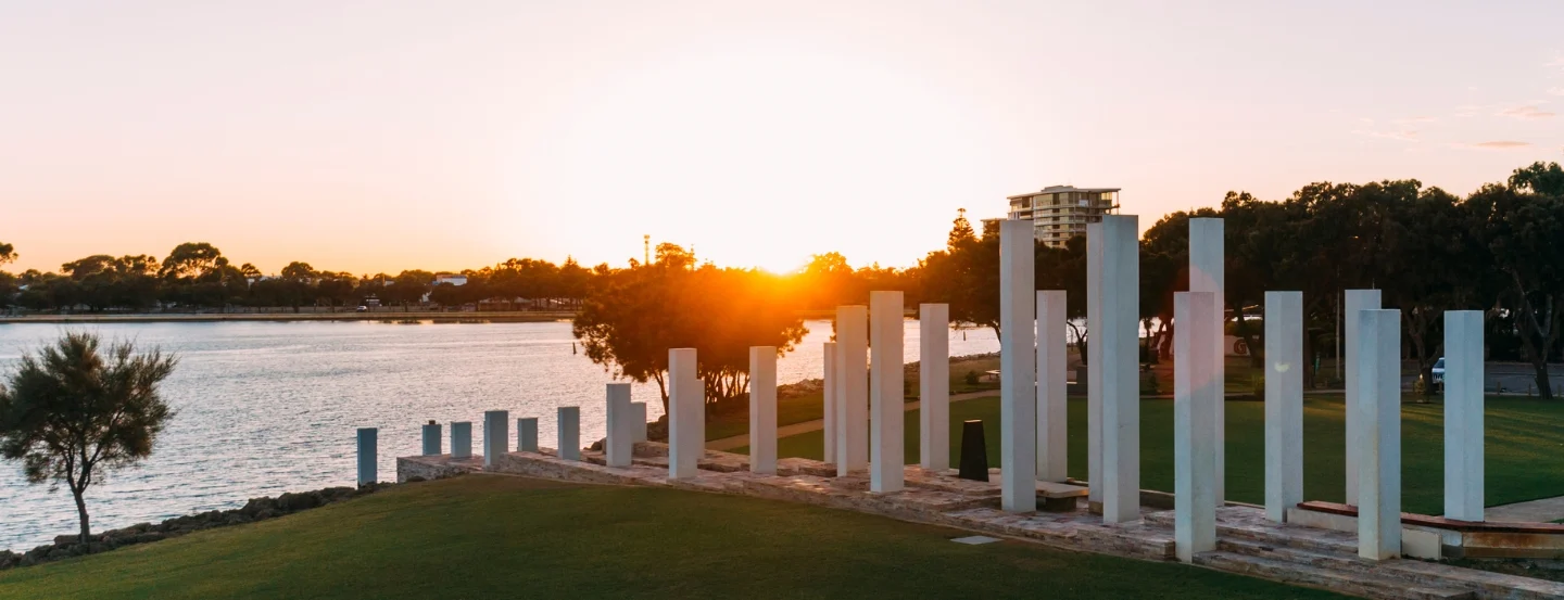 A scenic view of a grassy riverbank at sunset, with tall, white rectangular columns standing in a row. The river is calm, reflecting the orange hue of the sunset. Trees and buildings are visible in the distance.