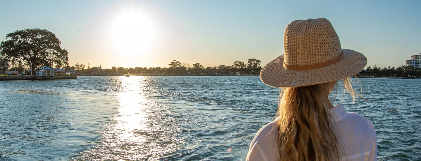 A woman with long hair and a straw hat gazes at the sun setting over a serene body of water. Trees and buildings line the distant shore, and the sunlight reflects off the gentle waves.