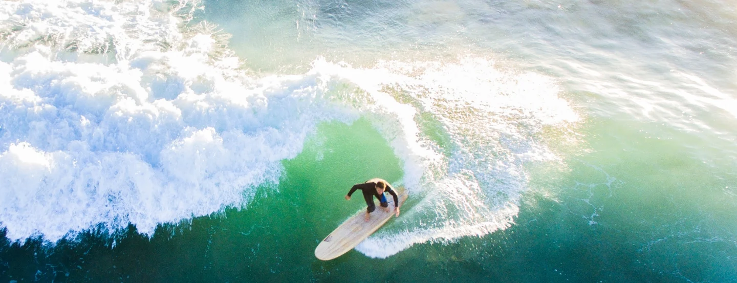 Aerial view of a surfer riding a wave on a longboard in clear turquoise water. The wave is cresting behind, creating a trail of white foam. Sunlight reflects on the water, highlighting the surfers skillful turn.
