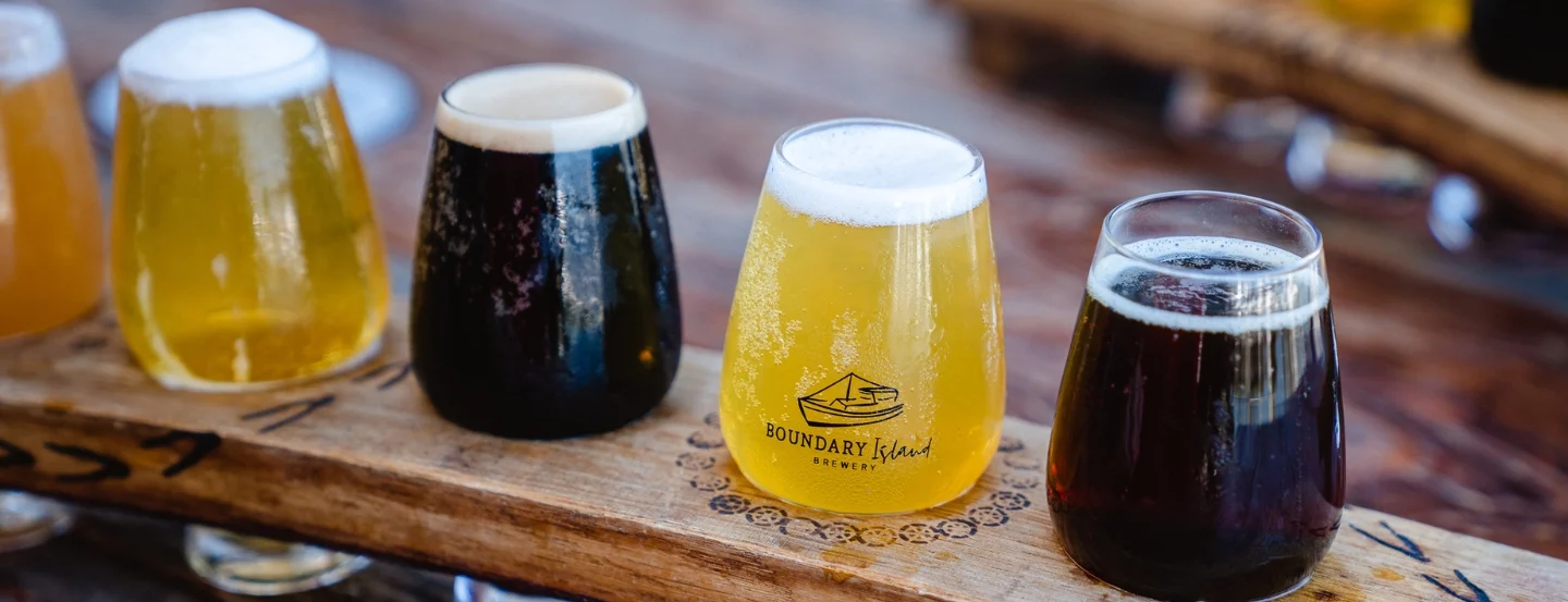 A wooden flight board holding four different beers, varying in color from light to dark. Each glass is filled with a frothy head. The beer in the center has Boundary Island Brewery printed on it. The board is set on a wooden table.