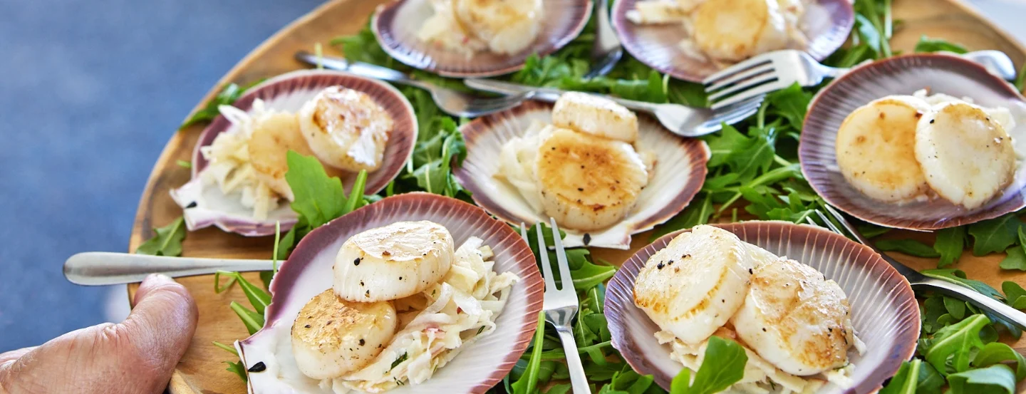 A person is holding a platter with scallop shells filled with seared scallops and coleslaw, garnished with arugula. Several small forks are placed beside the shells on the wooden tray.