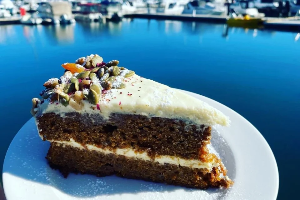 A slice of carrot cake with cream cheese frosting and pumpkin seed topping sits on a white plate. The background features a blurred view of a marina with boats and blue water.