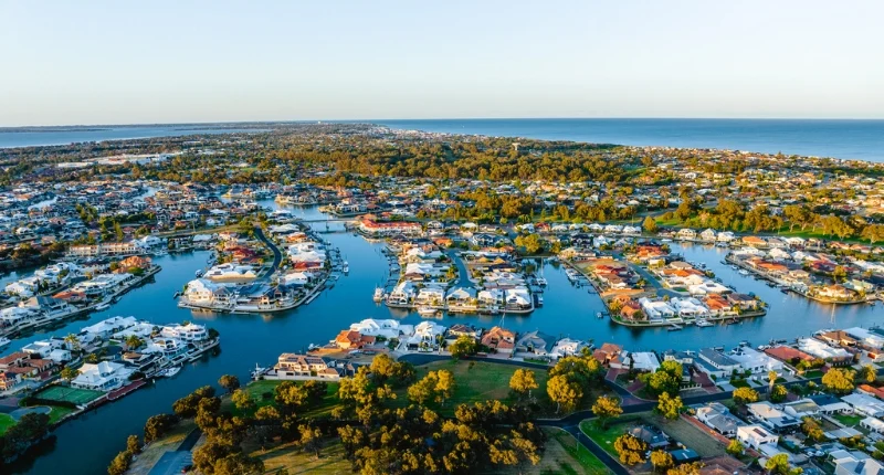 Aerial view of a coastal suburb featuring numerous houses lined along winding waterways, leading to a vast ocean. The foreground has green areas and trees, with a clear blue sky overhead.