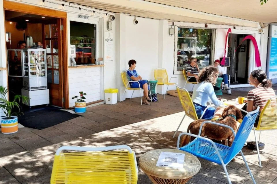 Outdoor seating at a cafe with people sitting on colorful chairs, some with dogs. The entrance is open, displaying a menu and plants. A tree casts shadows on the ground, creating a relaxed, sunny atmosphere.
