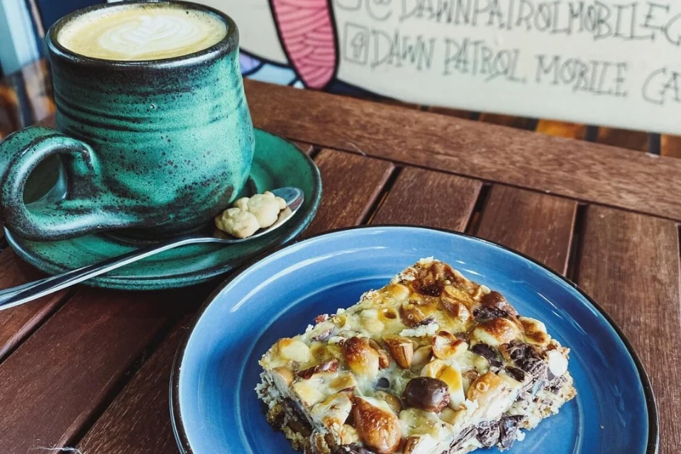 A green mug of latte on a wooden table next to a blue plate holding a dessert bar with marshmallows and nuts. A spoon with a small cream is beside the mug. The background features a colorful wall with text.