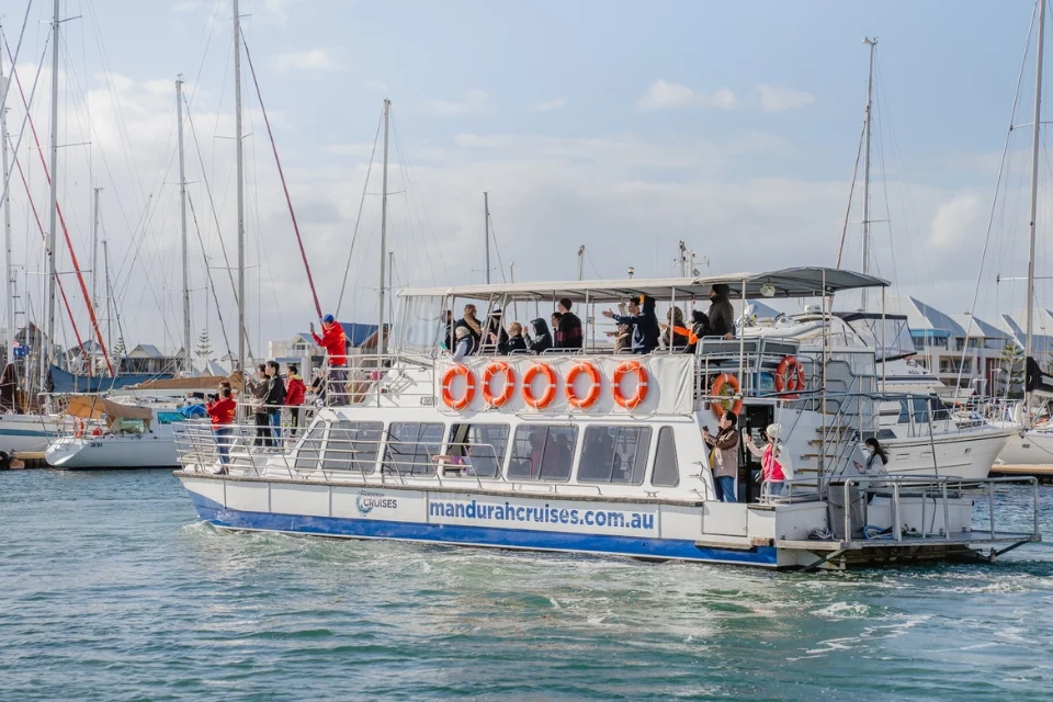 Tour boat in a marina with people on board, some taking photos. The boat has life rings and a website link on its side. Sailboats and masts are visible in the background under a partly cloudy sky.
