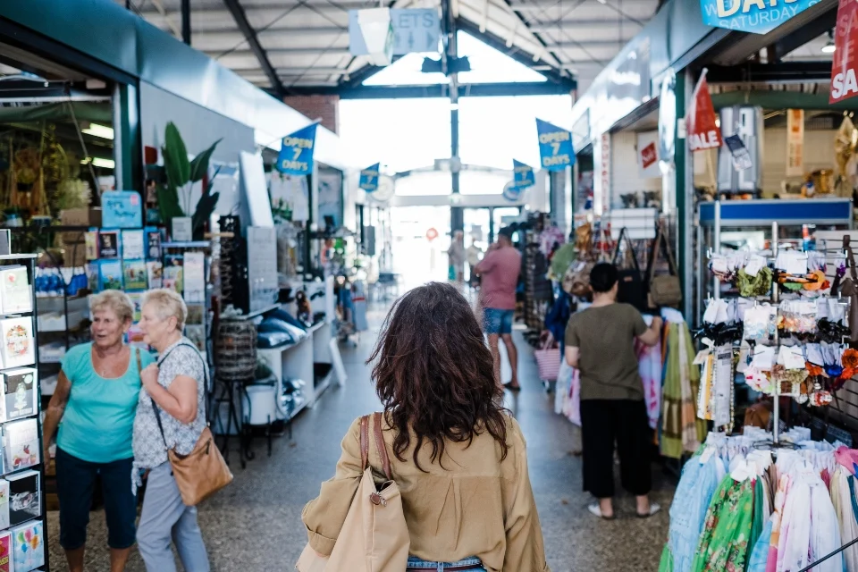 A woman with wavy hair walks through a bustling indoor market. People are shopping around her, browsing various stalls displaying cards, clothing, and goods. Signs indicate sales and store hours. Natural light fills the space from large windows above.
