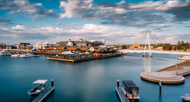 A marina with several boats docked, surrounded by water. There is a large building with a pier in the center and a cable-stayed pedestrian bridge to the right. The sky is partly cloudy, casting reflections on the calm water.