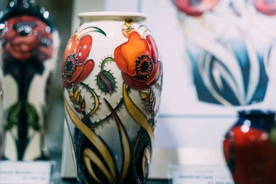 A decorative vase with vibrant red poppy flowers and intricate green leaves displayed in a shop. Other vases with similar floral designs can be seen in the background.