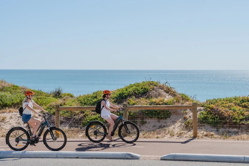 Two people wearing orange helmets ride bicycles along a paved path by the ocean. Low sand dunes with greenery are in the background under a clear blue sky.