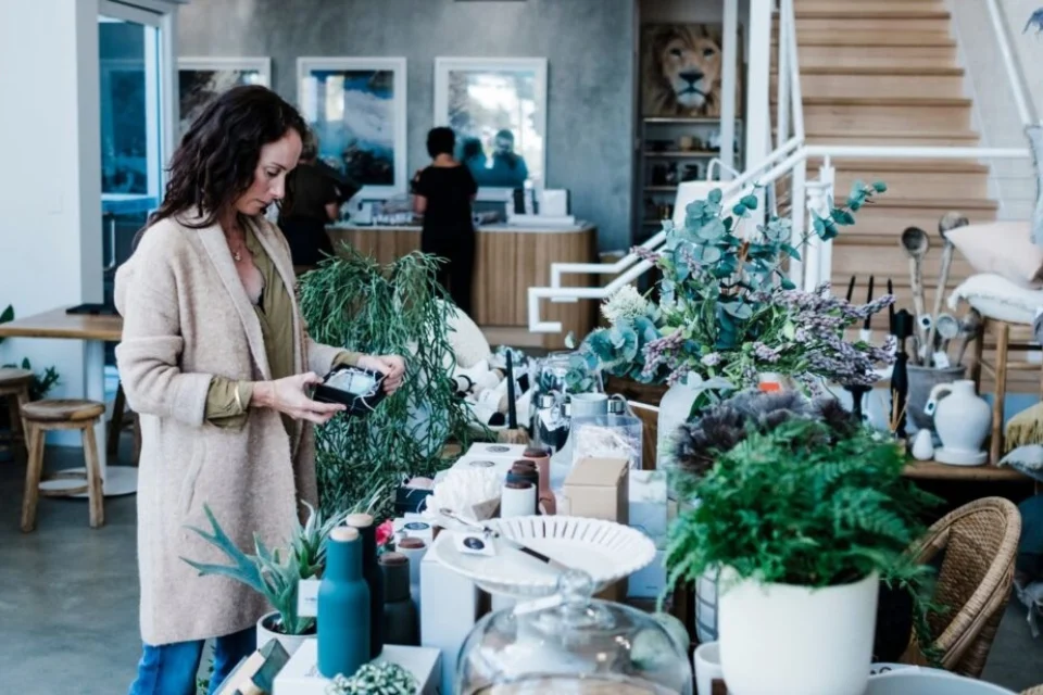 
A woman in a beige coat browses various items on a table in a store. The table displays plants, bottles, and decorative objects. Two people are at the counter in the background near a wall with framed animal portraits and a staircase.
