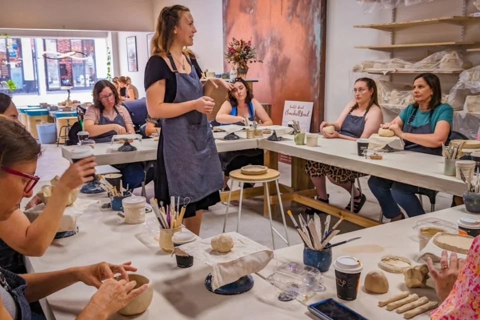 A pottery class with a female instructor guiding a group of adults seated around tables. The tables are filled with clay, tools, brushes, and cups. Shelves with pottery supplies are in the background. Participants listen and shape their clay.