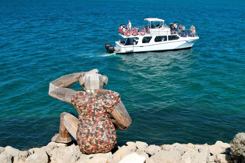 A large wooden sculpture resembling a human figure sits on rocks, facing a motorboat on a calm blue sea. The sculpture appears to be observing the boat, which is filled with people enjoying the ride under a clear sky.