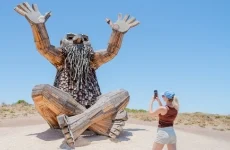A woman wearing a blue cap and shorts takes a photo with her phone of a large, whimsical wooden sculpture of a bearded figure sitting cross-legged with arms raised under a clear blue sky.