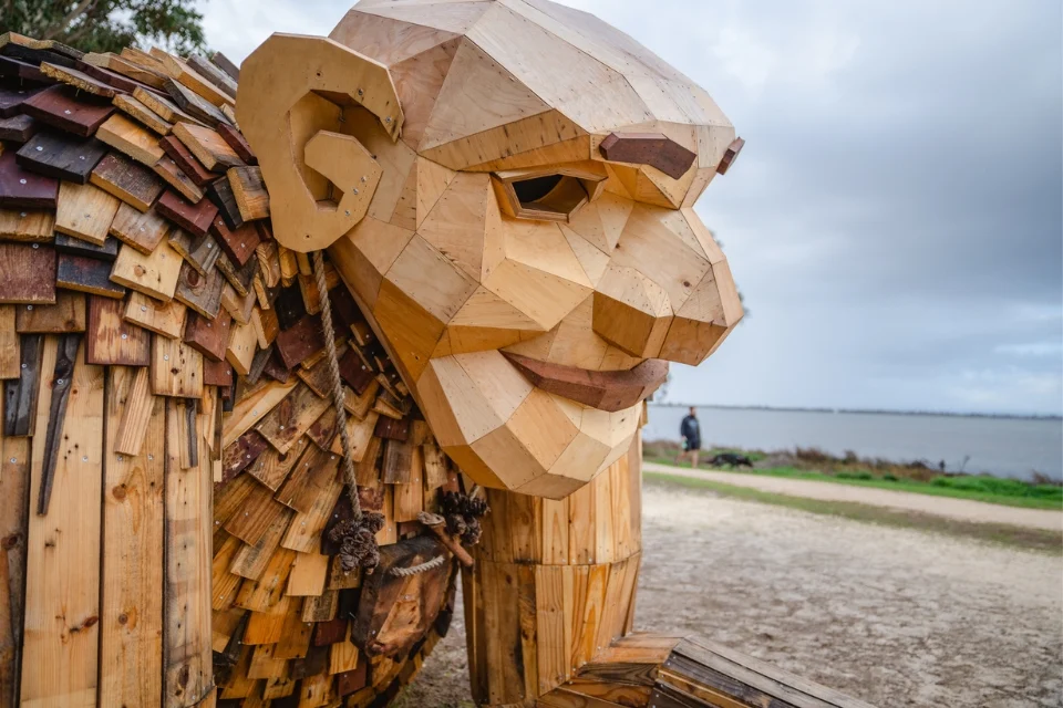 A large wooden sculpture of a troll, constructed from various pieces of light and dark wood, sits near a gravel path by the Mandurah estuary. The sky is overcast, and theres a grassy area in the background.