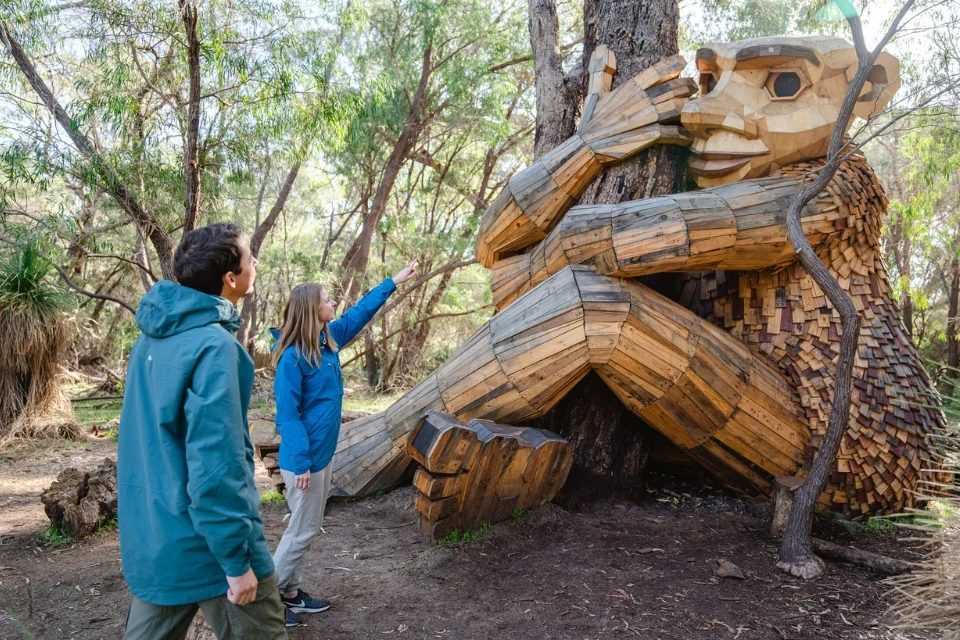 Two people in outdoor clothing admire a large wooden sculpture of a sitting figure with one hand over its eyes, leaning against a tree. The artwork is surrounded by lush greenery and trees.