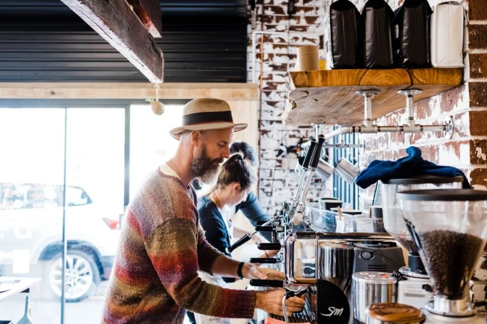 A man in a hat and sweater prepares coffee at an espresso machine in a rustic café. A woman works alongside him. Shelves with coffee bags are above them, and a window shows a parked car outside.