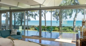 Living room with glass doors leading to a wooden deck overlooking the ocean. Two turquoise chairs are on the deck. Indoor space features a blue sofa, a white cushion, and decor. Lush greenery is visible outside.