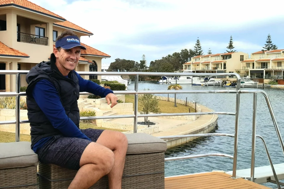 Justin Langer, former Australian cricket coach, in a cap and shorts sits on a boat. Behind him, there are boats, water, and several waterfront houses with terracotta roofs. The sky is cloudy, and trees line the background.