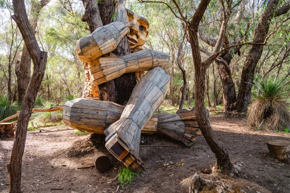 Wooden sculpture of a large, seated figure with a thoughtful pose, nestled among trees in a forest setting. The piece is creatively crafted from plank-like segments, blending with the natural surroundings.