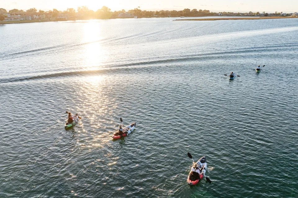 A group of people kayaking on a calm, wide river as the sun rises. The water reflects the golden light. In the background, there is a distant shoreline with buildings and trees silhouetted against the sky.