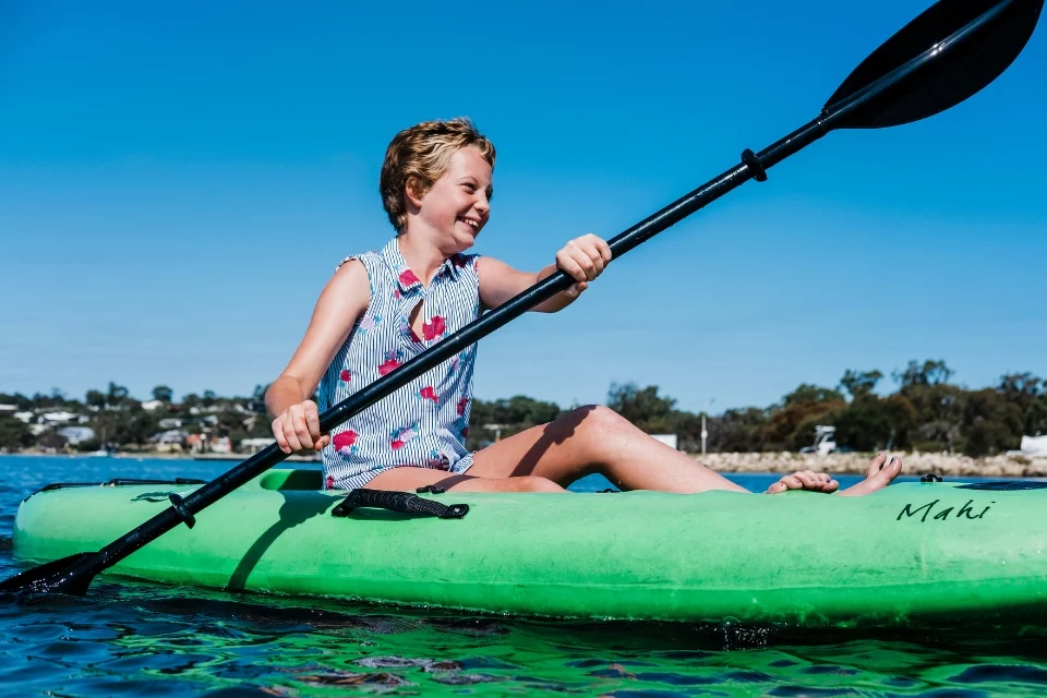 A young girl is smiling while kayaking on calm blue water in a bright green kayak. She is wearing a sleeveless shirt with a blue and white pattern and red flowers. The background shows trees and a few buildings under a clear blue sky.