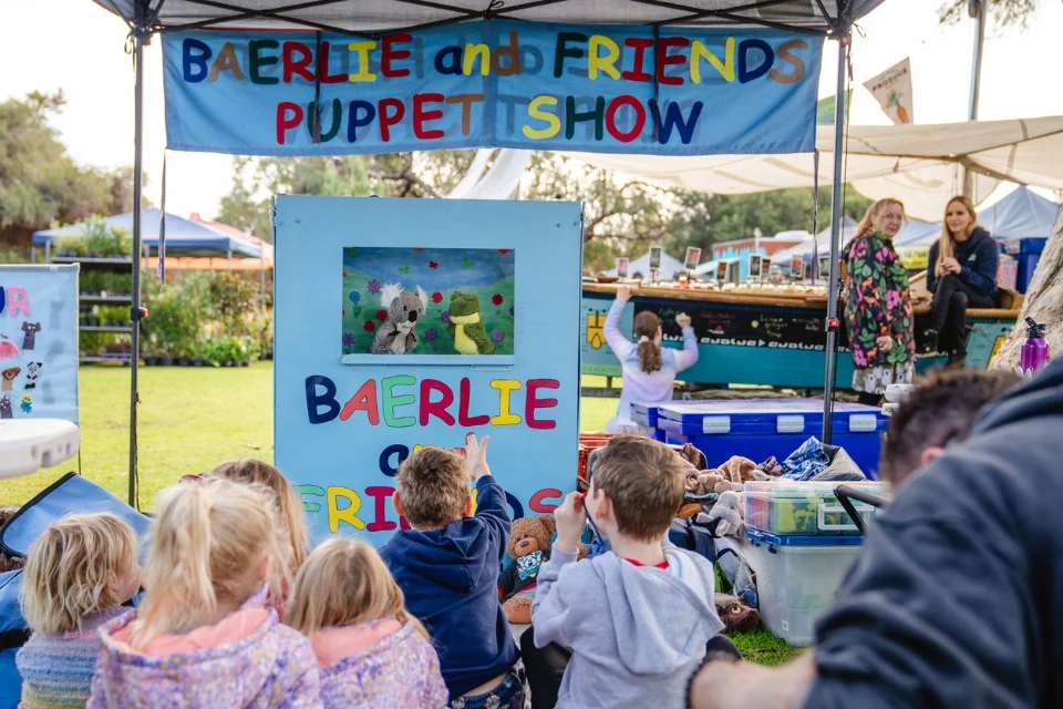 Children seated outdoors watching a puppet show titled Baerlie and Friends. A colorful banner decorates the stage. A lively crowd is gathered, including adults and kids enjoying the sunny day at what appears to be a community event or fair.