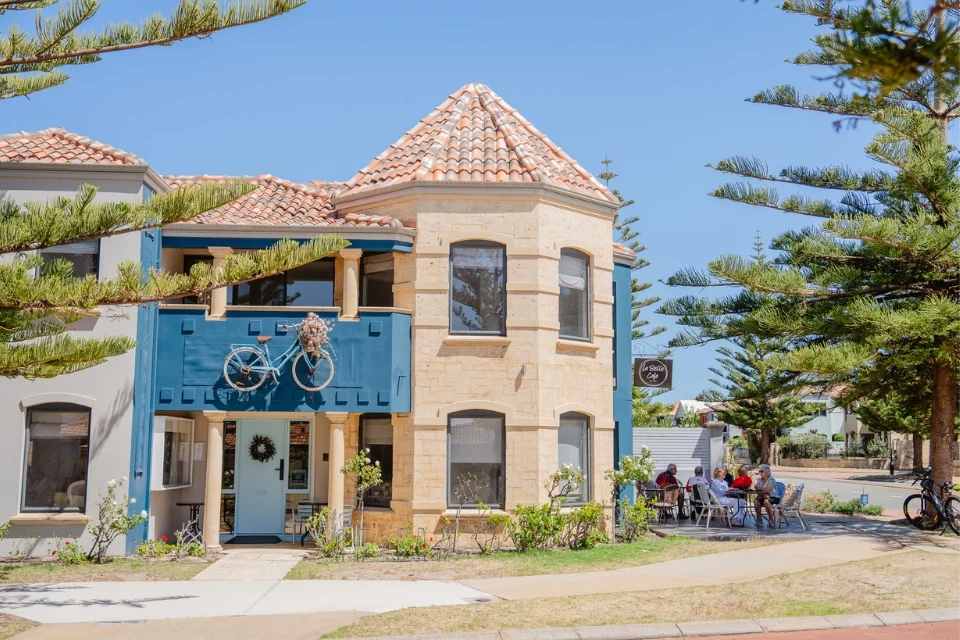 A quaint two-story building with a mix of blue and beige stone exterior features a bicycle mounted above the entrance. Outdoor seating with people can be seen to the right under tall pine trees. The sky is clear and blue.