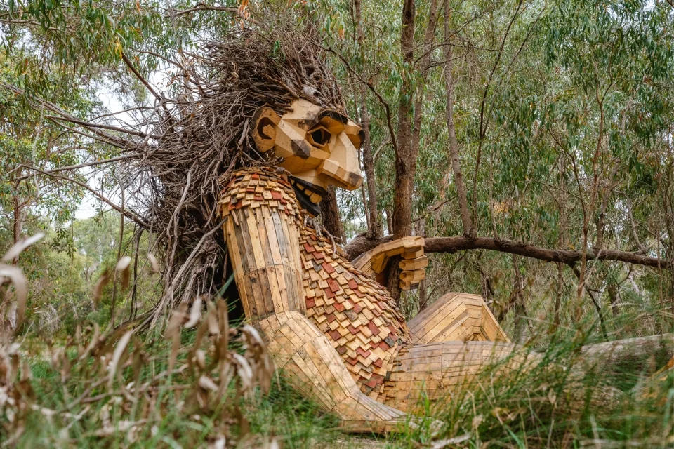 A large wooden sculpture of a mythical creature with a human-like form and leafy hair sits nestled among trees, holding a stick. The art piece is crafted from various shades of wood, blending naturally into the forest setting.