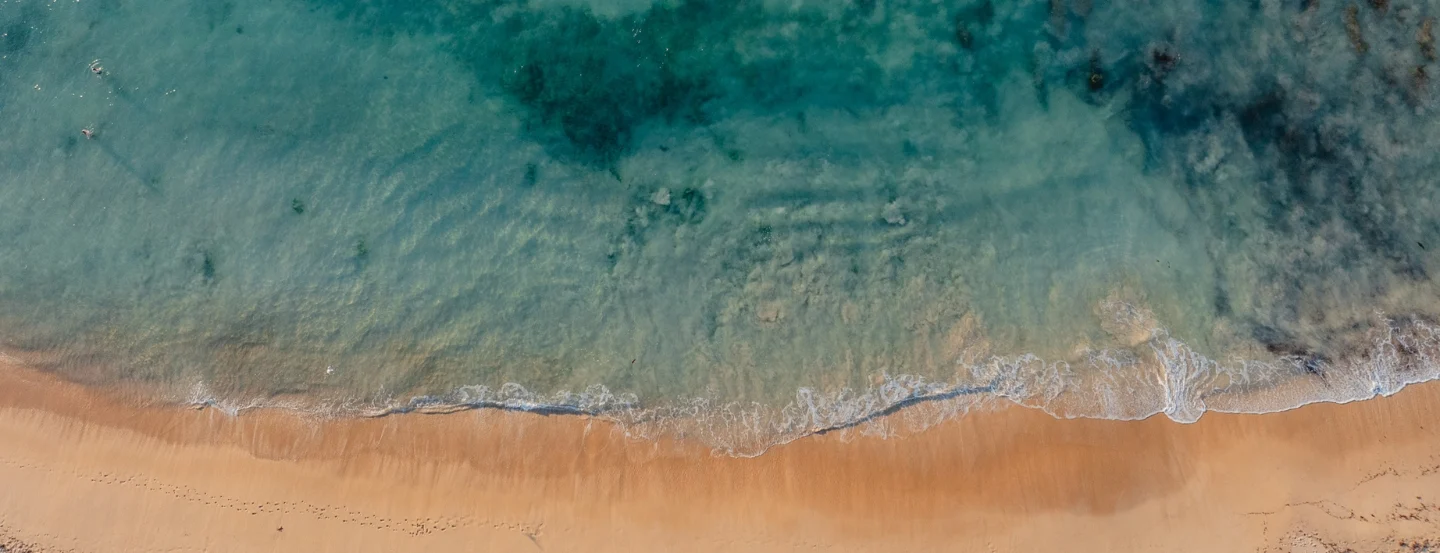 Aerial view of a sandy beach with gentle ocean waves washing onto the shore, and clear blue-green water fading into deeper shades.