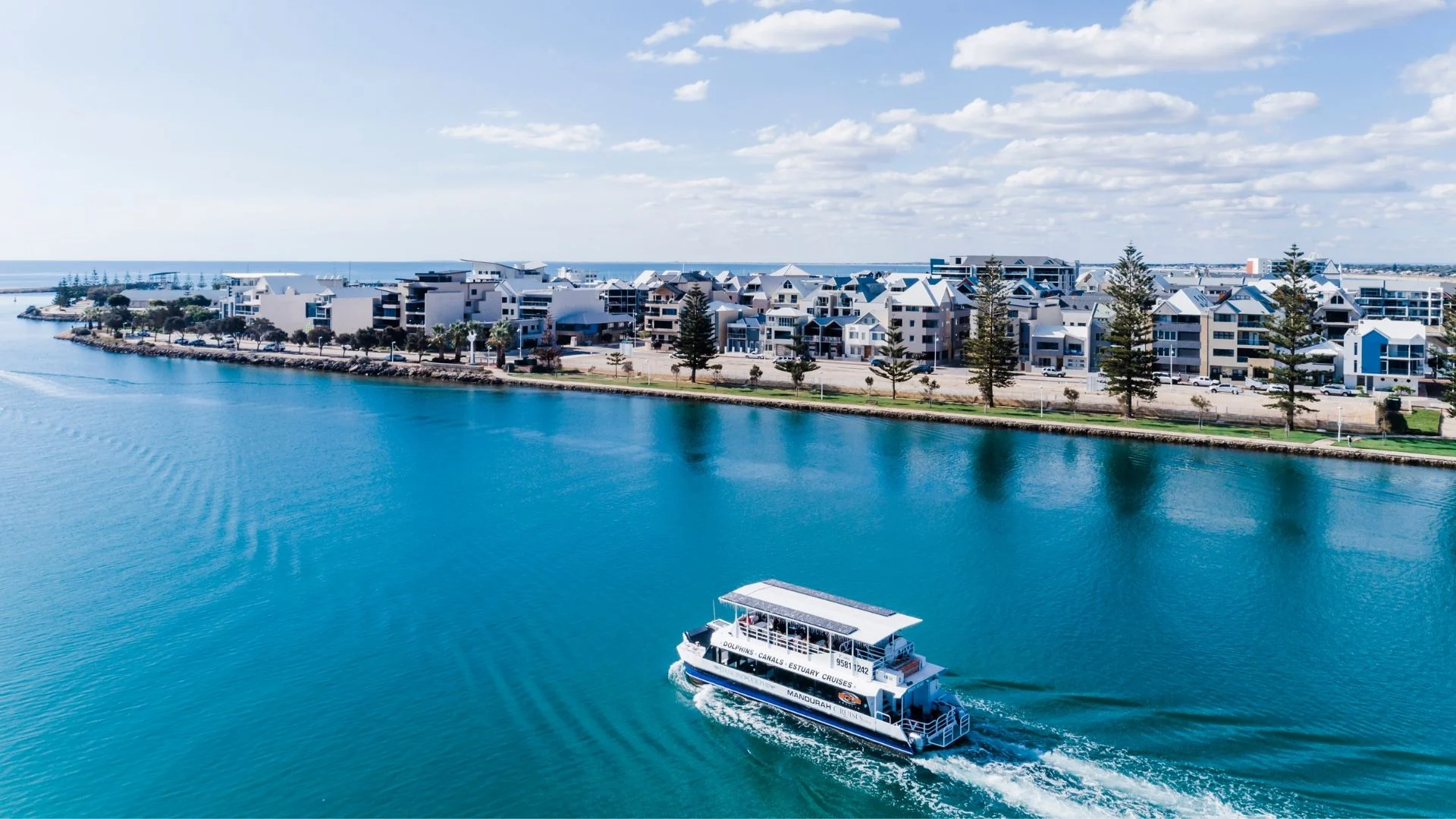 Aerial view of a boat navigating a wide, calm blue river. On the right, there are modern white buildings with a row of trees in front. The sky is partly cloudy, adding to a serene coastal atmosphere.