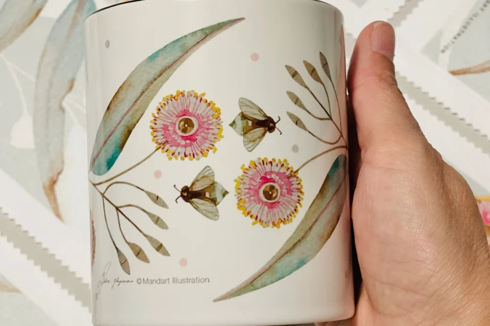 A hand holds a white ceramic mug with a botanical illustration featuring pink and yellow flowers, two bees, and green leaves. The background is a pattern of paper-like textures with leaf designs.
