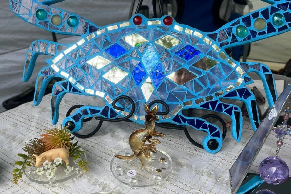 A table displays a mosaic art piece shaped like a crab with blue and silver tiles. In front, there are small glass figurines, including a pig and a dinosaur, each set on separate bases.