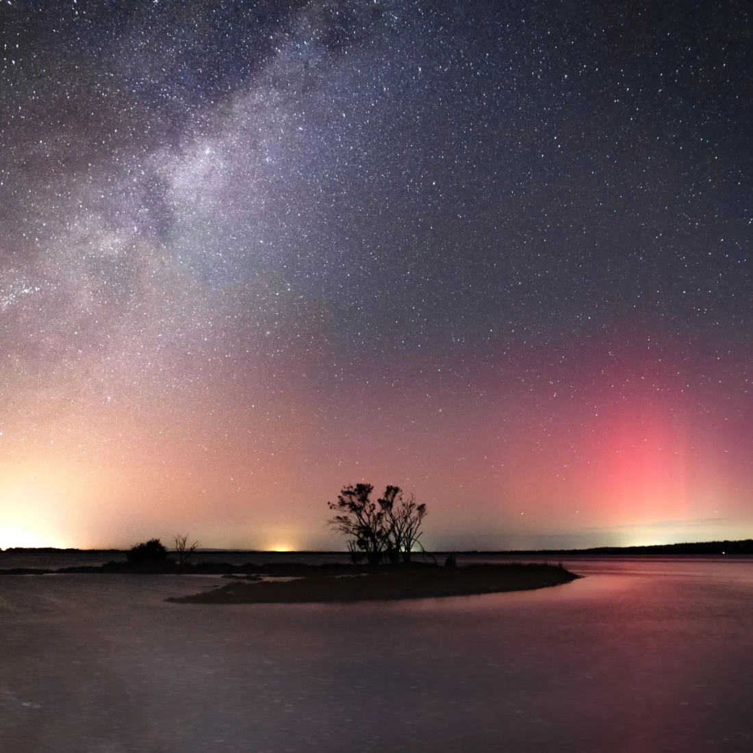 A starry night sky with the Milky Way and colourful auroras glows above a calm lake, silhouetting trees on a small island. The water reflects the lights, creating a peaceful, otherworldly scene.