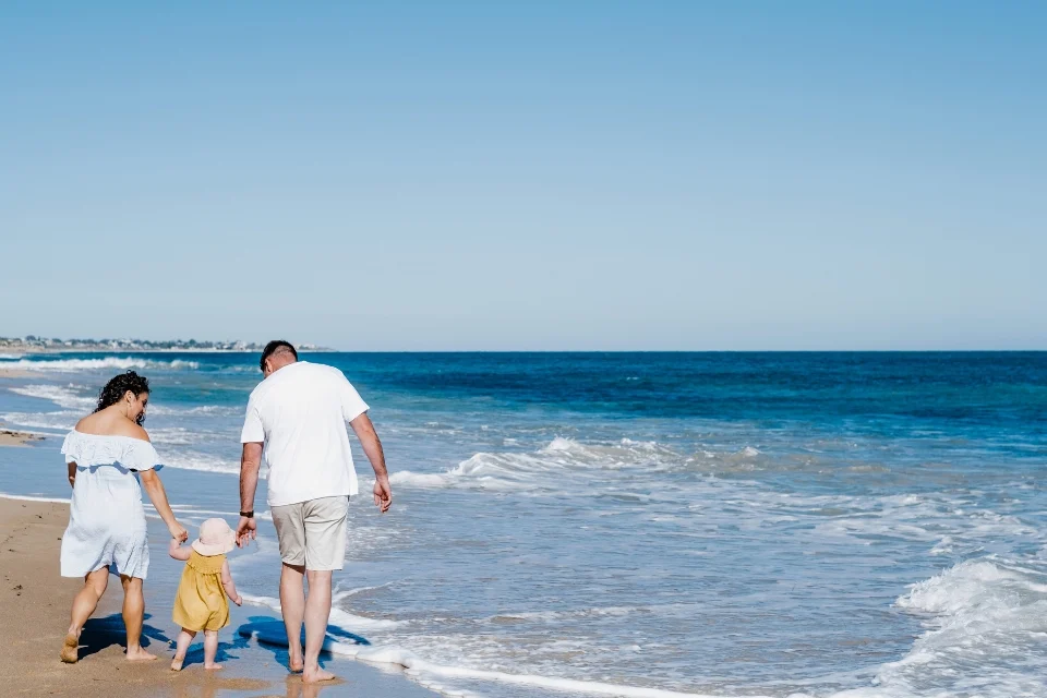 A family of three walks barefoot along a sunny beach. The child, wearing a yellow outfit and sunhat, holds hands with two adults. The ocean waves gently wash onto the shore, and the sky above is clear and blue.