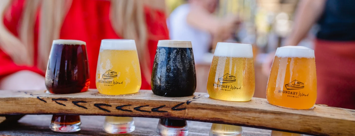 A wooden paddle holds five small glasses of different colored beers, ranging from dark to light. A person in a red shirt is partially visible in the background, sitting at a table outdoors.