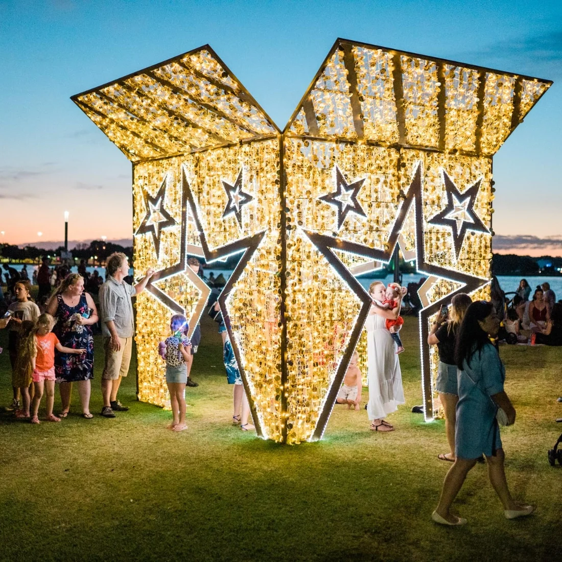 People gather around and inside a large, illuminated gift box structure decorated with stars on a grassy area near a body of water at dusk. Families and children enjoy the festive, glowing display.