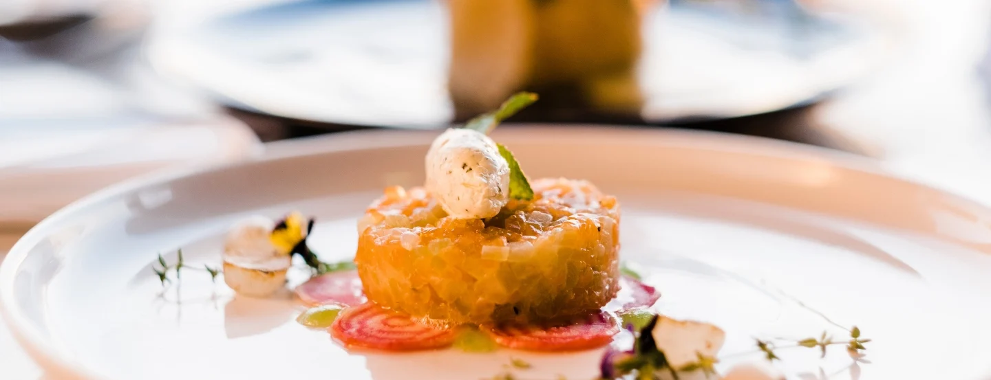 A gourmet dish is elegantly presented on a white plate, featuring a round portion of finely chopped vegetables topped with a dollop of cream. Thinly sliced vegetables and edible flowers garnish the plate, adding vibrant colour.