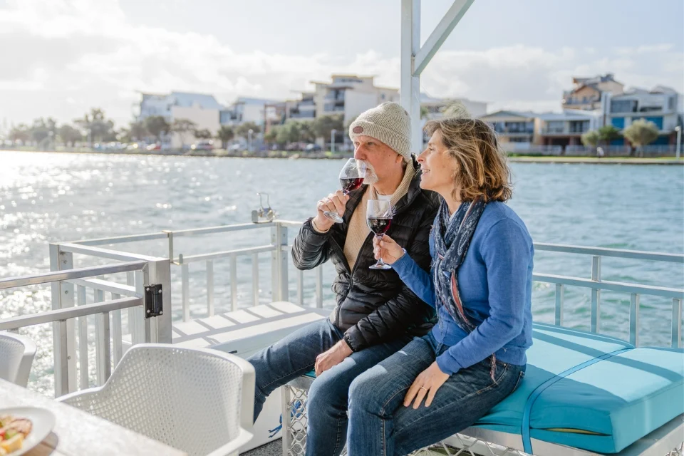 A man and woman sit on a boat by the water, holding glasses of red wine. The man wears a hat and jacket, while the woman is dressed in a blue sweater and scarf. Buildings line the shore in the background under a partly cloudy sky.