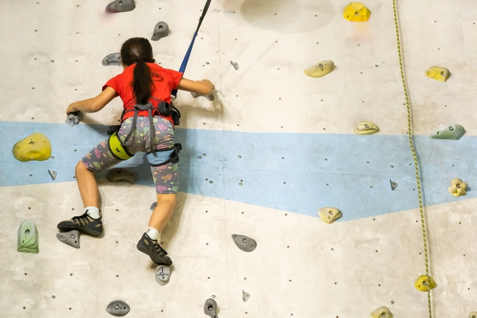 A person wearing a red shirt and colourful leggings climbs an indoor rock wall with a safety harness. They are reaching for holds on the light-colored wall, which has various colored grips and a blue stripe running diagonally.