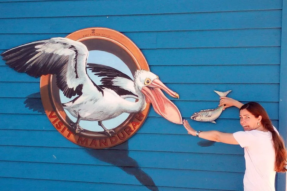 A woman playfully holds a fish in front of a 3D wall mural depicting a pelican with its beak open, appearing to reach for the fish. The mural is framed by a circular border with the words Visit Mandurah. The background is a vibrant blue wall.