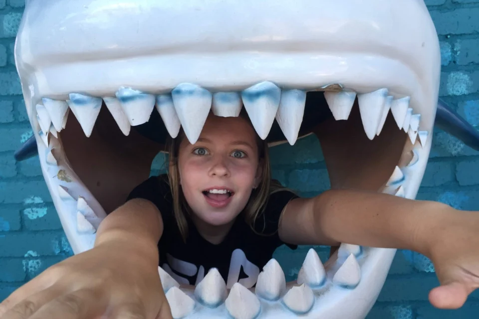 A child playfully poses inside a large shark mouth display with wide open jaws. The child reaches forward with a playful expression against a blue brick background.