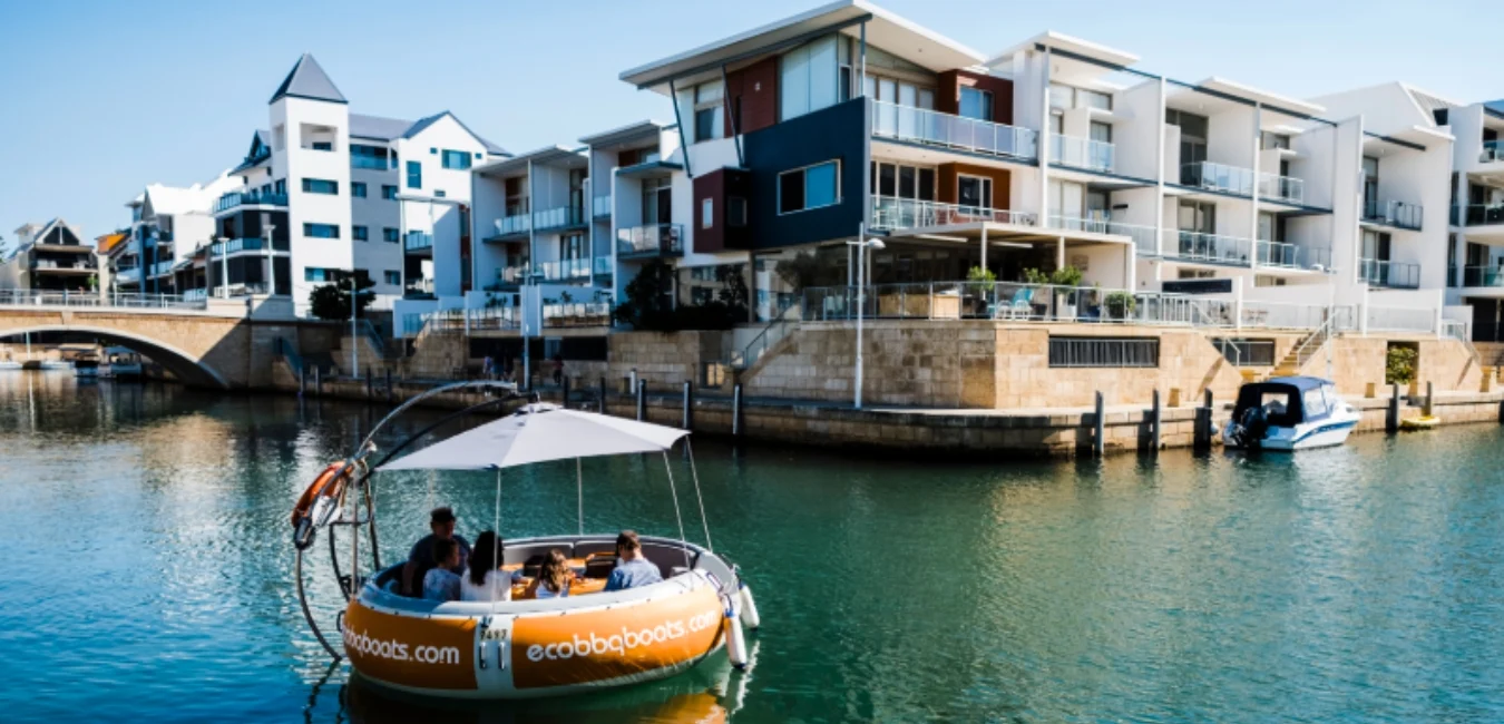 A floating, round boat with a canopy carrying a group of people is docked by modern waterfront apartments. The scene is sunny, showcasing a calm blue canal with a small bridge in the background.